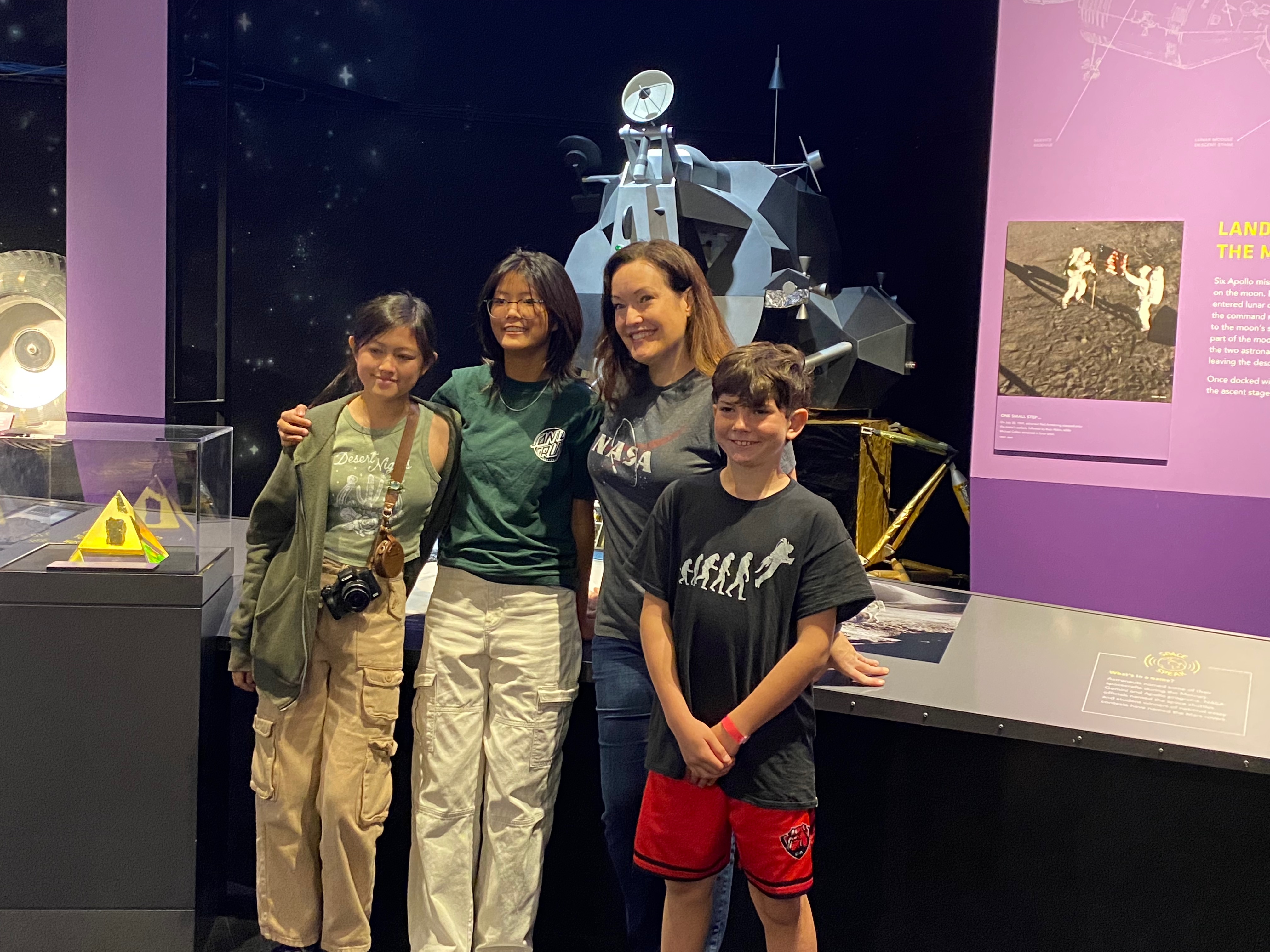 Three kids and a woman stand close together with big smiles in front of a museum display.